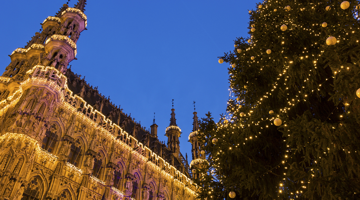 A tall Christmas tree decorated in gold lights and baubles next to Leuven City Hall which is illuminated by lights